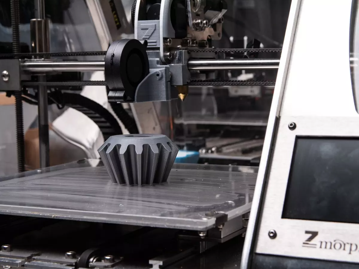 Close-up shot of a 3D printer working. The printer is printing a small gray object. The object is a gear with six points.