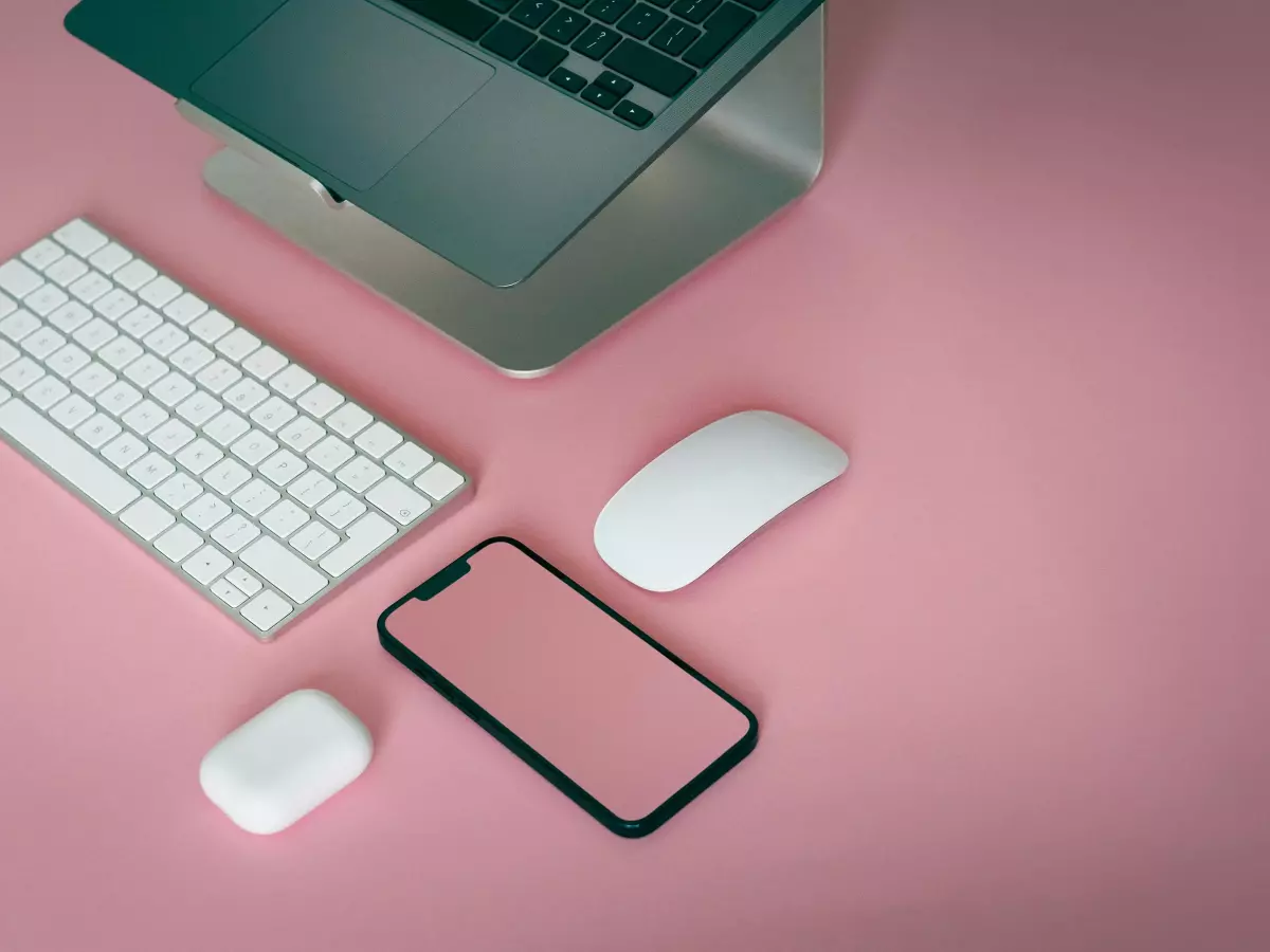 A smartphone, laptop, keyboard, and mouse on a pink surface, with the smartphone's screen showing a blank pink background.