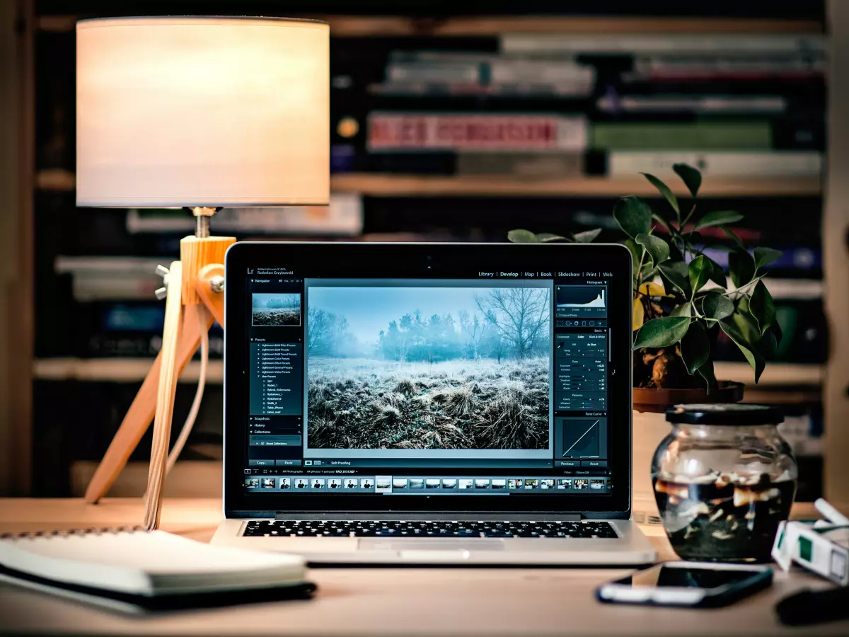 A laptop computer on a desk with a lamp in front of it, the laptop screen showing a snowy landscape and a digital photo editor open.