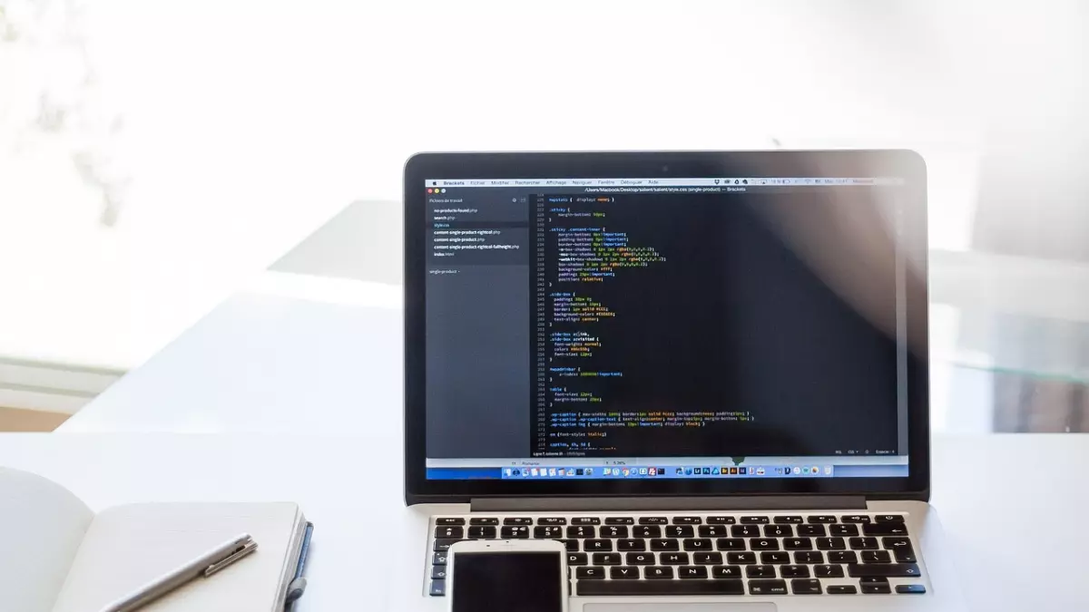 A laptop with code on the screen on a desk with a notebook and a phone.