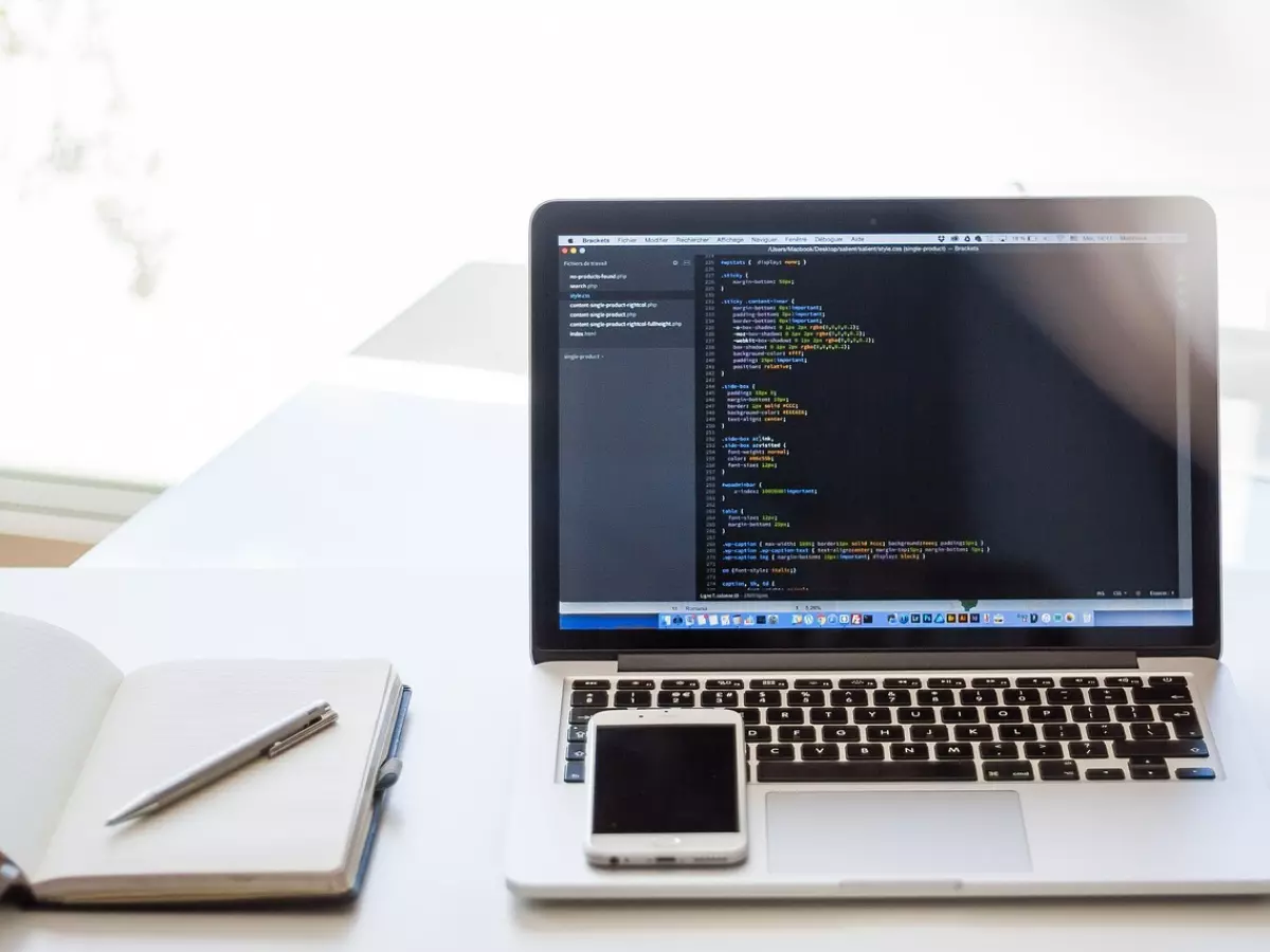 A laptop with code on the screen on a desk with a notebook and a phone.