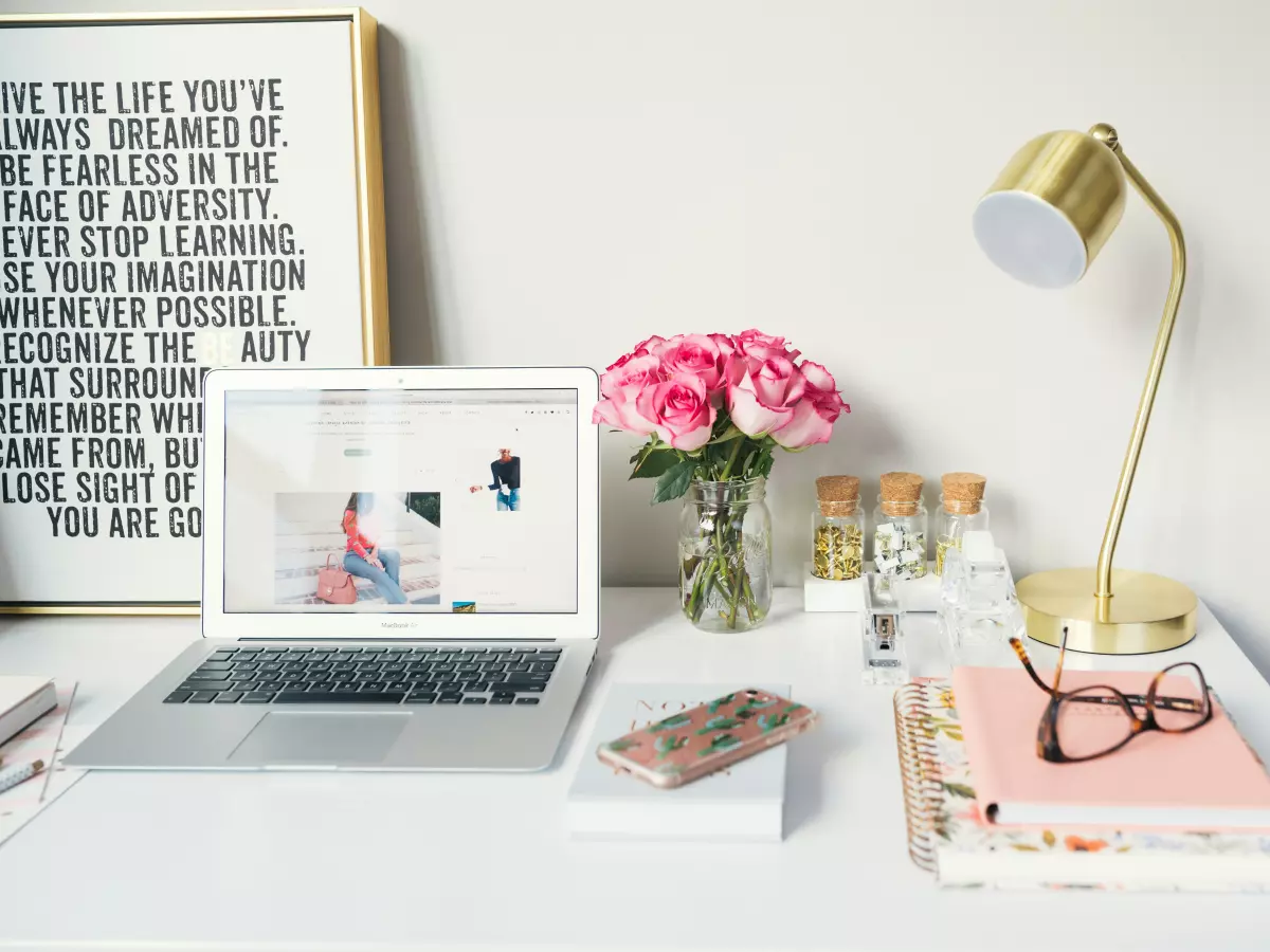 A white desk with a laptop, a lamp, flowers, and some books. The laptop is open and has a website open on it.