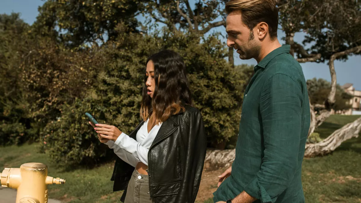 A woman in a white shirt and a black leather jacket is standing on a path in a park. She is looking at her phone, which is in her right hand. A man is standing behind her, looking at her. The man is wearing a green shirt and sunglasses.