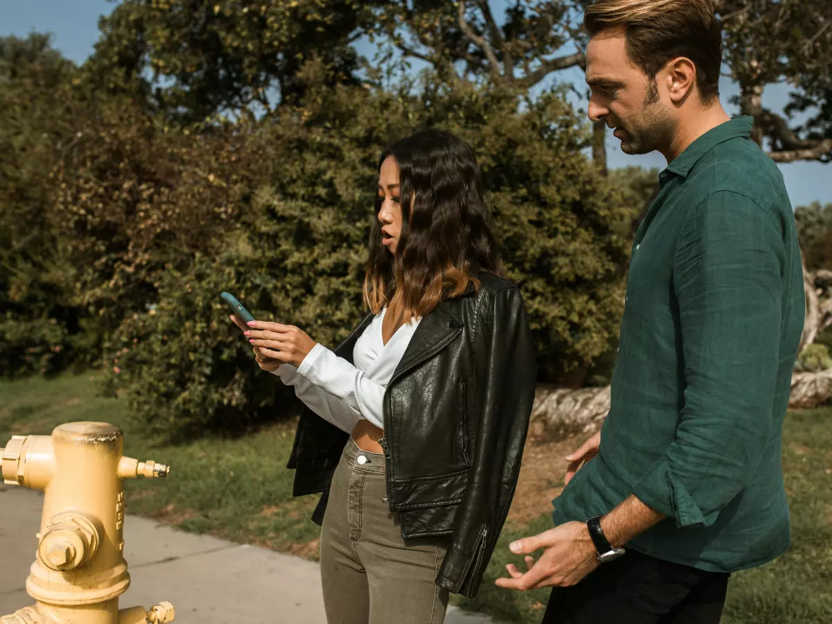 A woman in a white shirt and a black leather jacket is standing on a path in a park. She is looking at her phone, which is in her right hand. A man is standing behind her, looking at her. The man is wearing a green shirt and sunglasses.