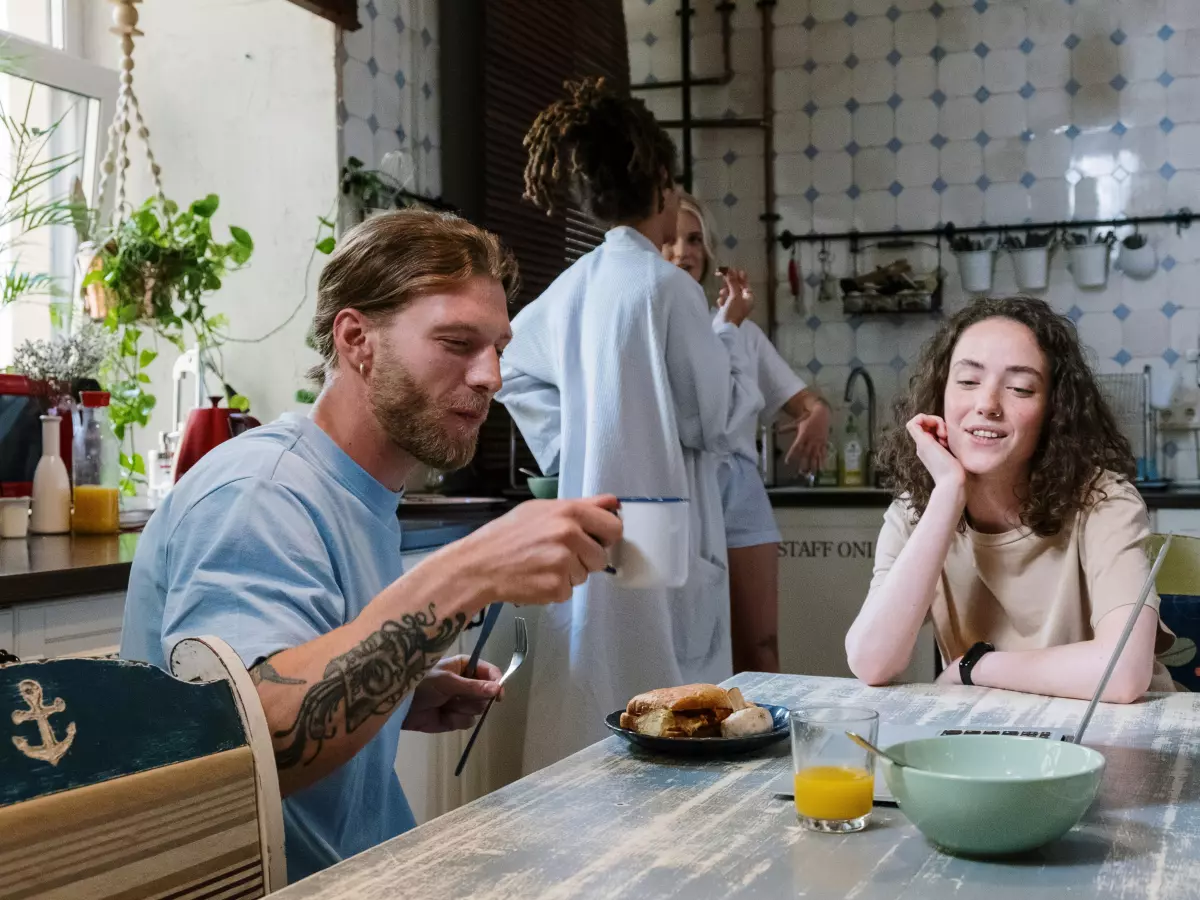 A man and a woman sitting at a kitchen table, the man is looking at his phone while the woman is looking at him. There is another person in the background.