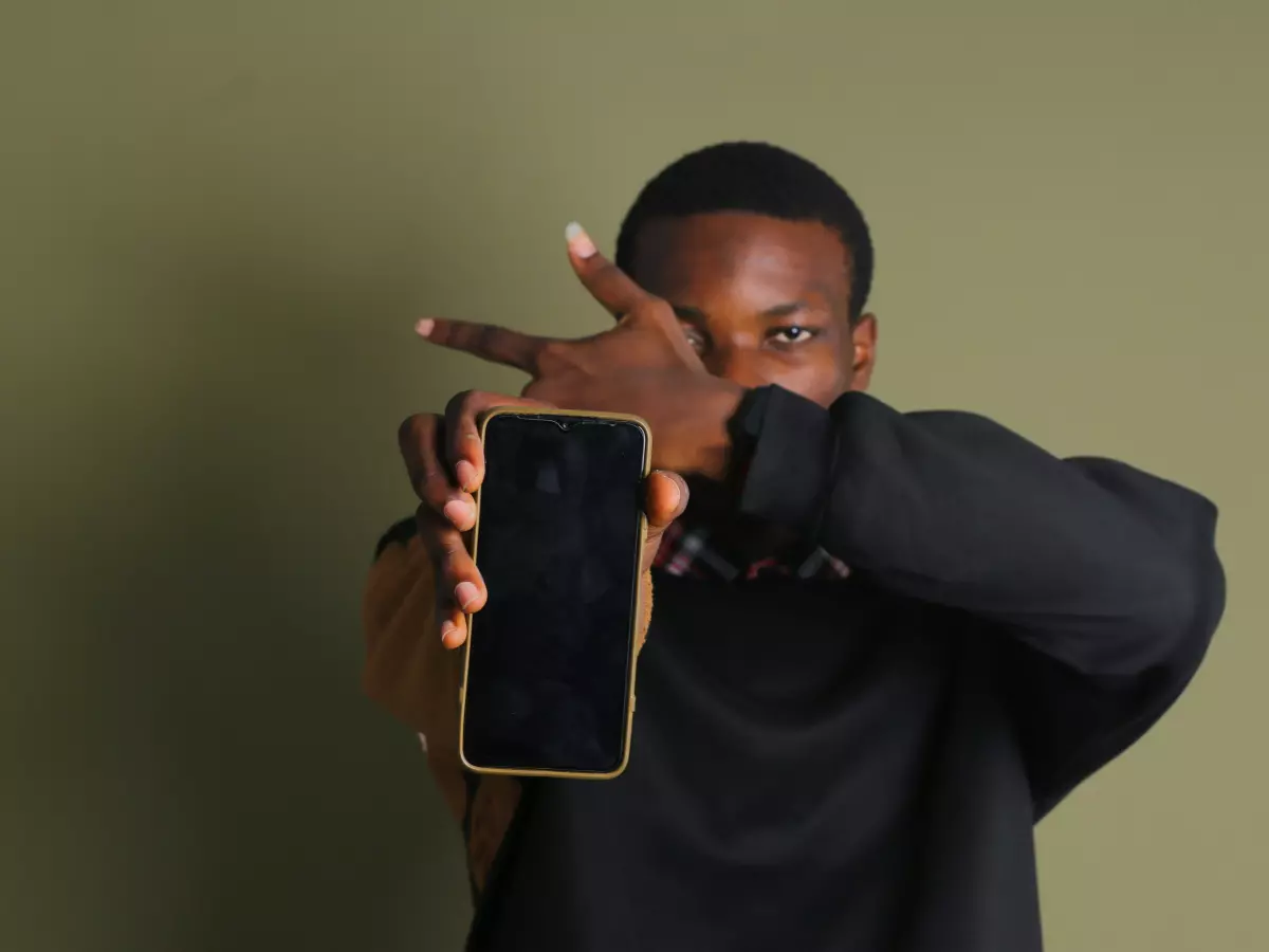 A young man is holding a smartphone in front of his face, with his hand covering his mouth and nose. The smartphone is showing a black screen.