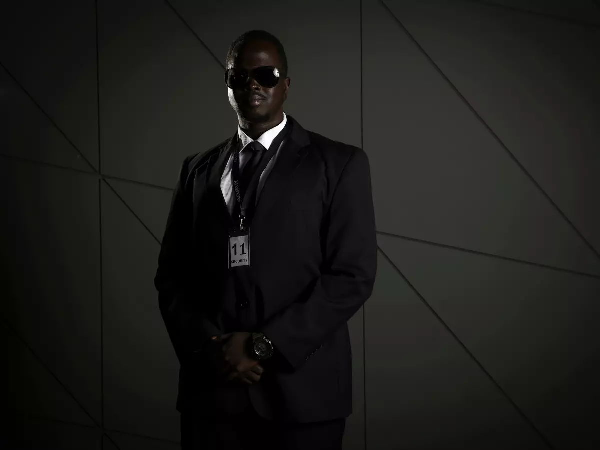 A security guard in a black suit and sunglasses standing in front of a grey wall.