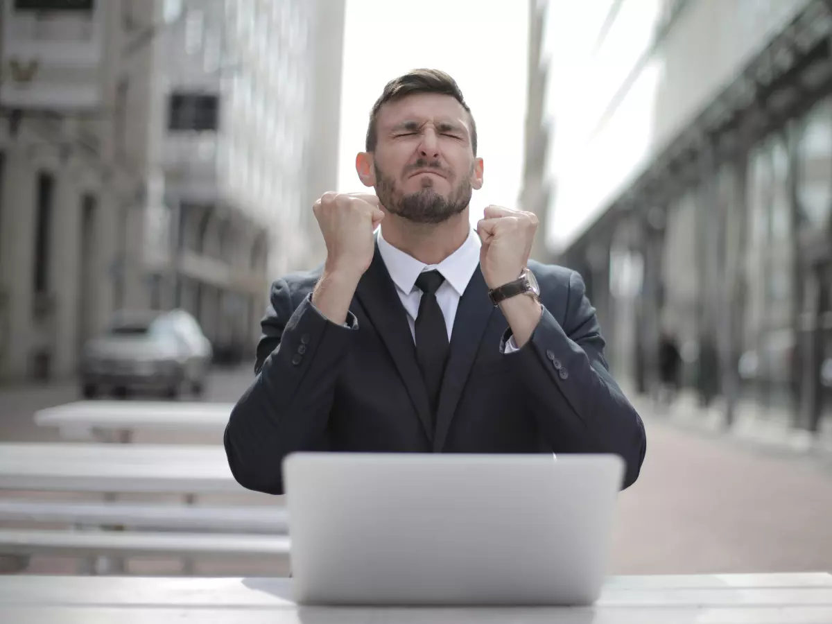 A businessman is sitting at a table outside with his laptop open. He is wearing a suit and tie. He is smiling and has his fists clenched in the air. There are buildings in the background and a car in the distance. The image is in a cool color palette of grays and blues.