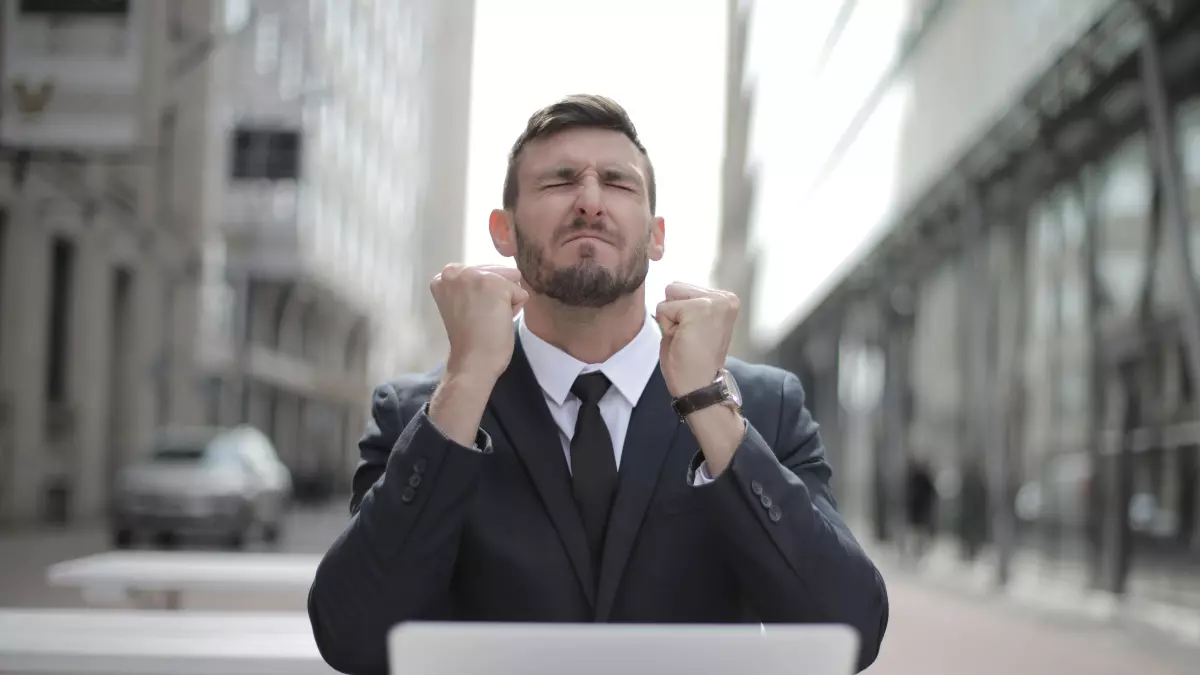 A businessman is sitting at a table outside with his laptop open. He is wearing a suit and tie. He is smiling and has his fists clenched in the air. There are buildings in the background and a car in the distance. The image is in a cool color palette of grays and blues.