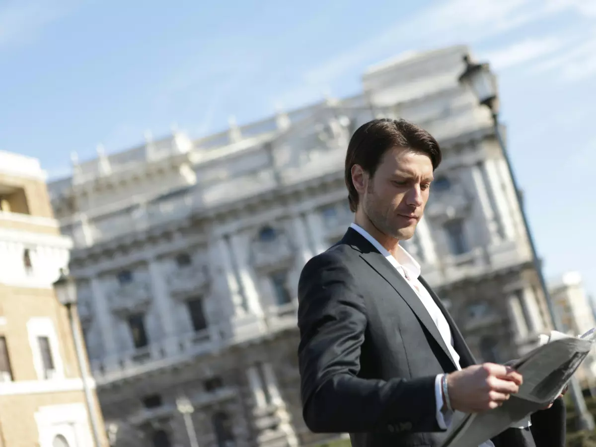 A man in a suit stands in front of a large building, holding a newspaper.