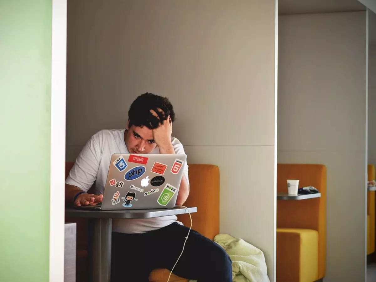 A young man is sitting at a table with a laptop, his hand on his head, looking distressed.