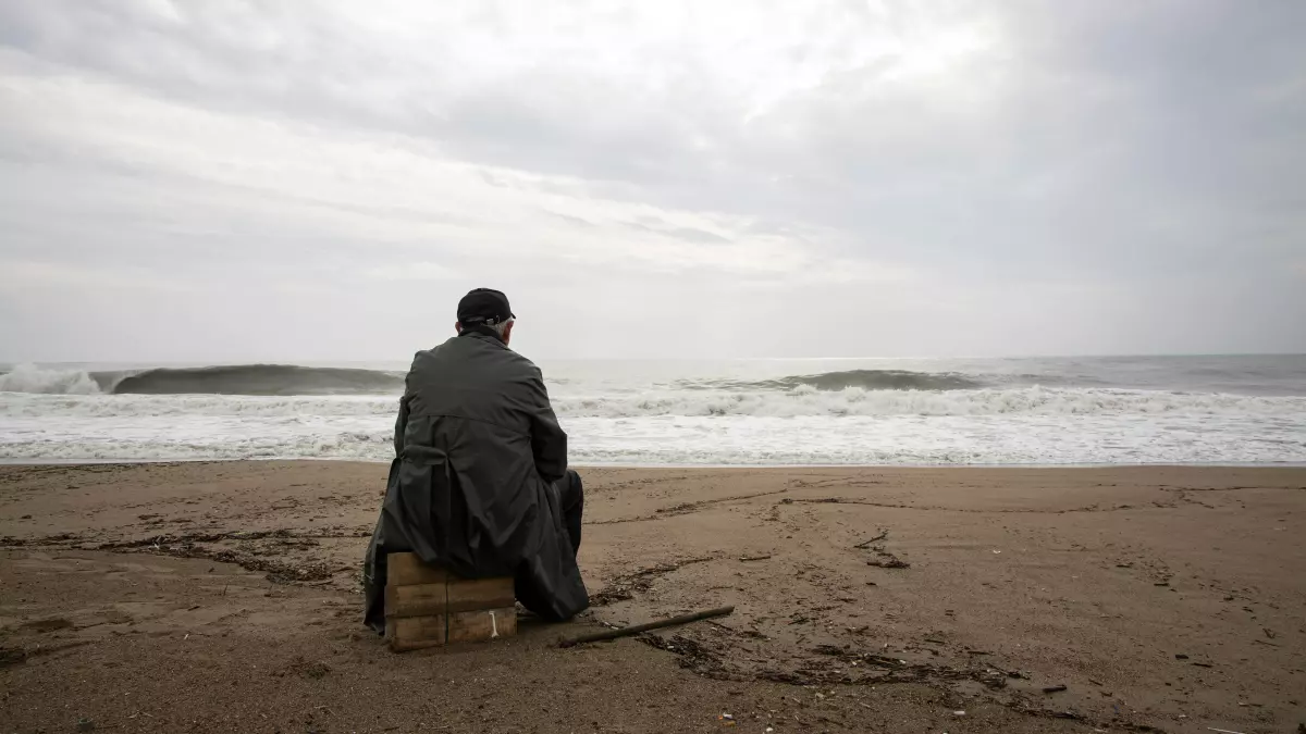 A man sits on a rock on a beach, looking out at the ocean. The sky is cloudy and the water is choppy.