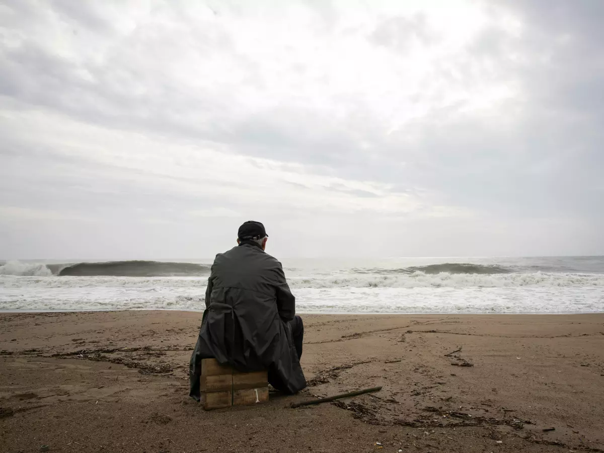 A man sits on a rock on a beach, looking out at the ocean. The sky is cloudy and the water is choppy.