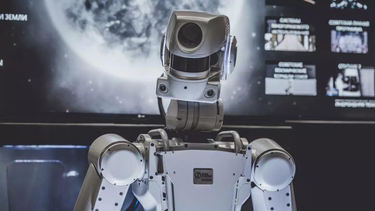 A white humanoid robot stands in front of a screen, its sensors and joints are visible. The robot is looking towards the viewer.