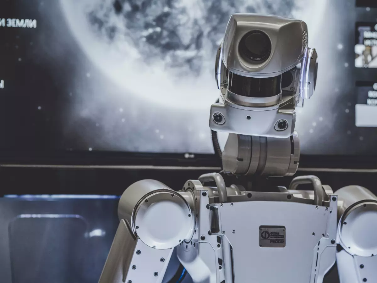 A white humanoid robot stands in front of a screen, its sensors and joints are visible. The robot is looking towards the viewer.