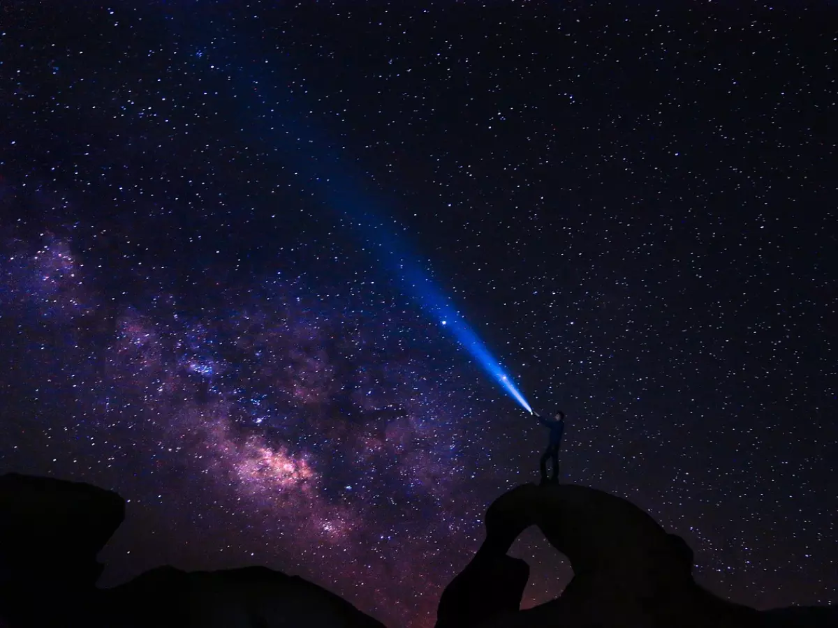 A silhouette of a person standing on a rock formation, shining a flashlight towards the Milky Way, which is visible in the night sky. The sky is full of stars, creating a breathtaking view.