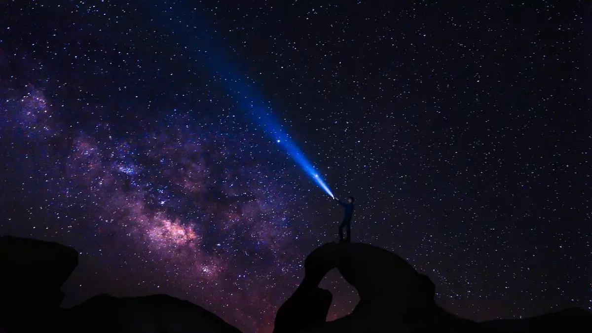 A silhouette of a person standing on a rock formation, shining a flashlight towards the Milky Way, which is visible in the night sky. The sky is full of stars, creating a breathtaking view.