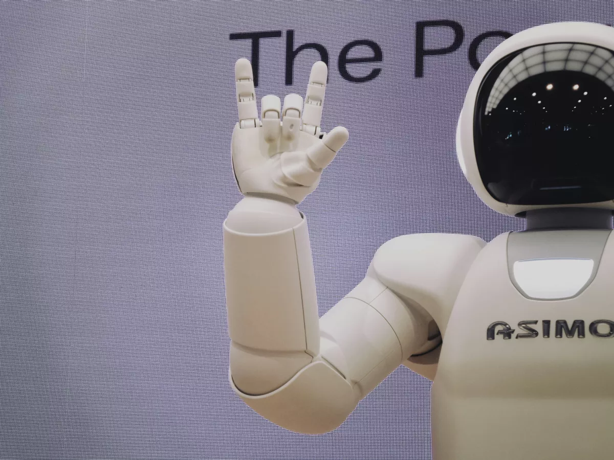 A humanoid robot with a white body and a black head, its right arm is raised, showing the fingers in a rock and roll gesture.