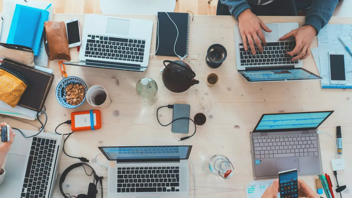 A top-down view of a table with multiple laptops, phones, and other electronic devices. People are working on the laptops and using the phones. 