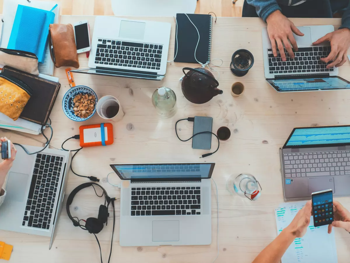 A top-down view of a table with multiple laptops, phones, and other electronic devices. People are working on the laptops and using the phones. 