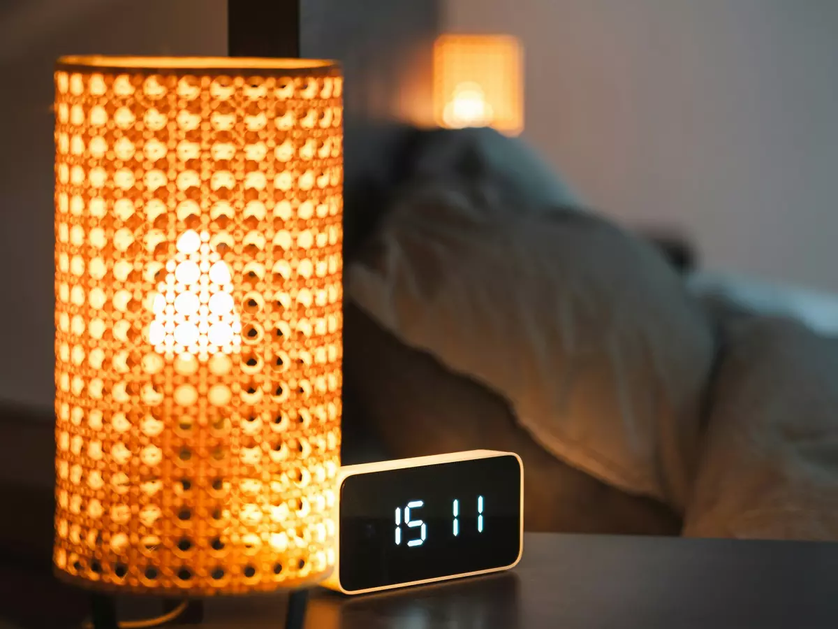 A close up image of a smart lamp on a night stand next to a digital clock, both are illuminated.