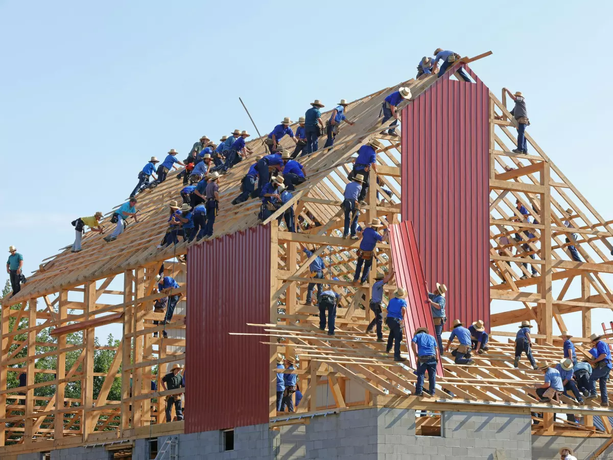 A group of people are working together to build a large wooden structure. The image is taken from a low angle, looking up at the structure. The people are wearing blue shirts and are working on the roof, walls, and frame of the structure.