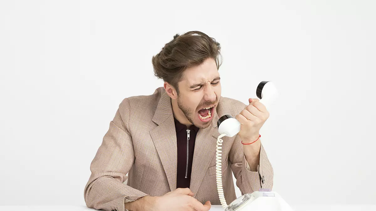 A man in a blazer is sitting at a desk with a phone in his hand, yelling angrily into the receiver.