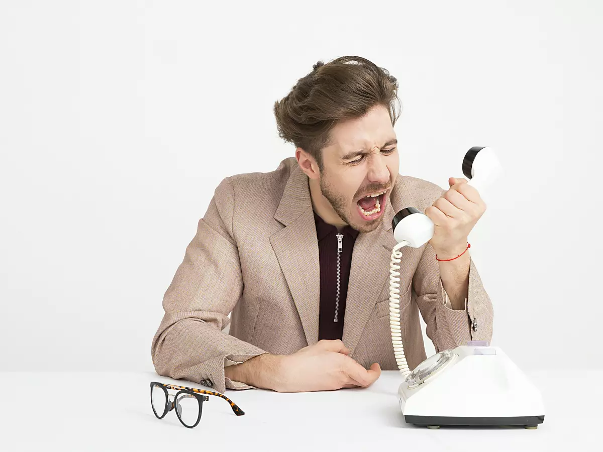 A man in a blazer is sitting at a desk with a phone in his hand, yelling angrily into the receiver.