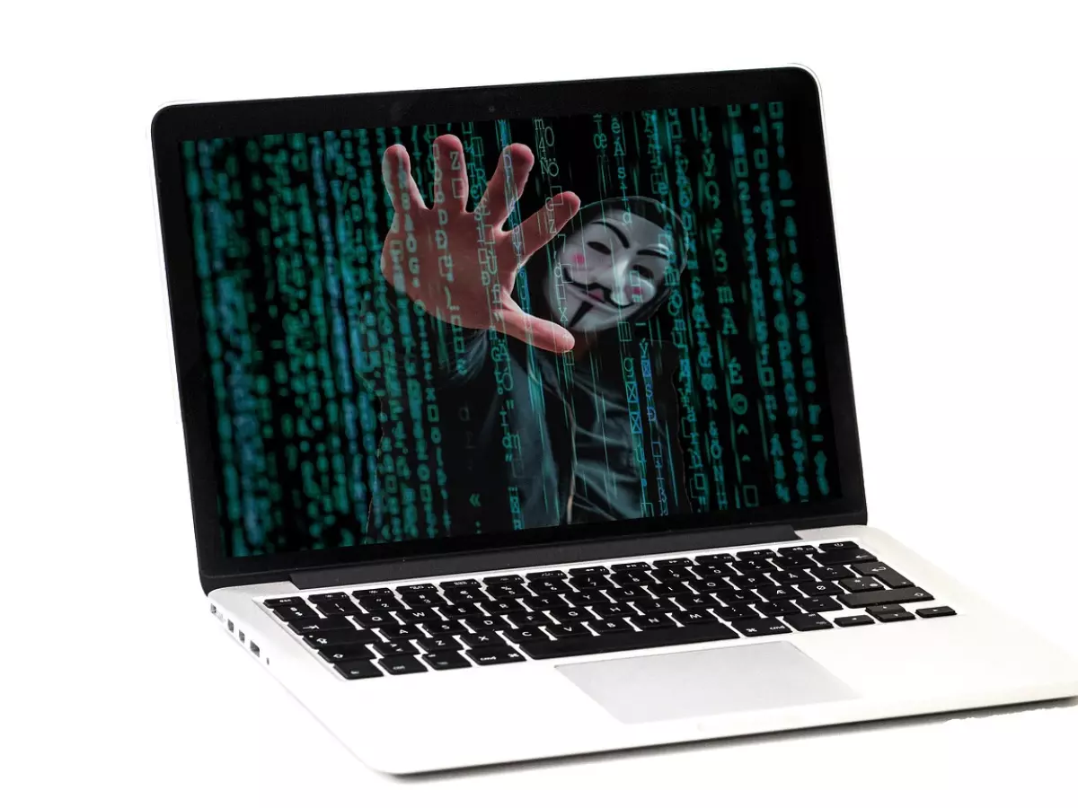 A hacker wearing a Guy Fawkes mask is reaching out of a laptop screen, which displays a digital code in the background.