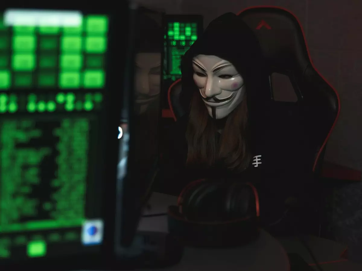 A person in a black hoodie with the face covered in a Guy Fawkes mask, is sitting in front of a computer, typing, looking serious.