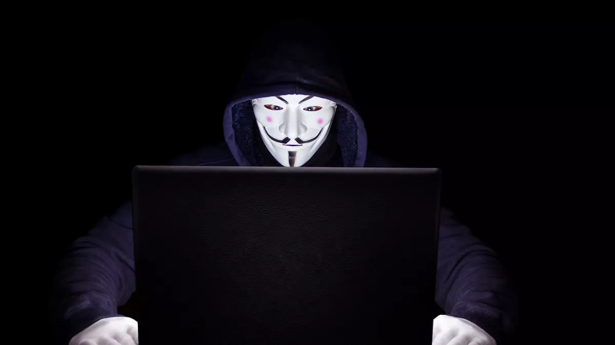 A person wearing a hooded sweatshirt and a Guy Fawkes mask is looking at a laptop. They have white gloves on. The background is dark.