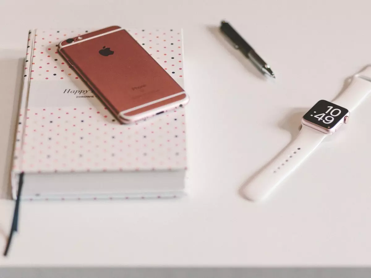 A rose gold iPhone and Apple watch are lying on a white table. There is a notebook, a pen, and a white watch band next to them.