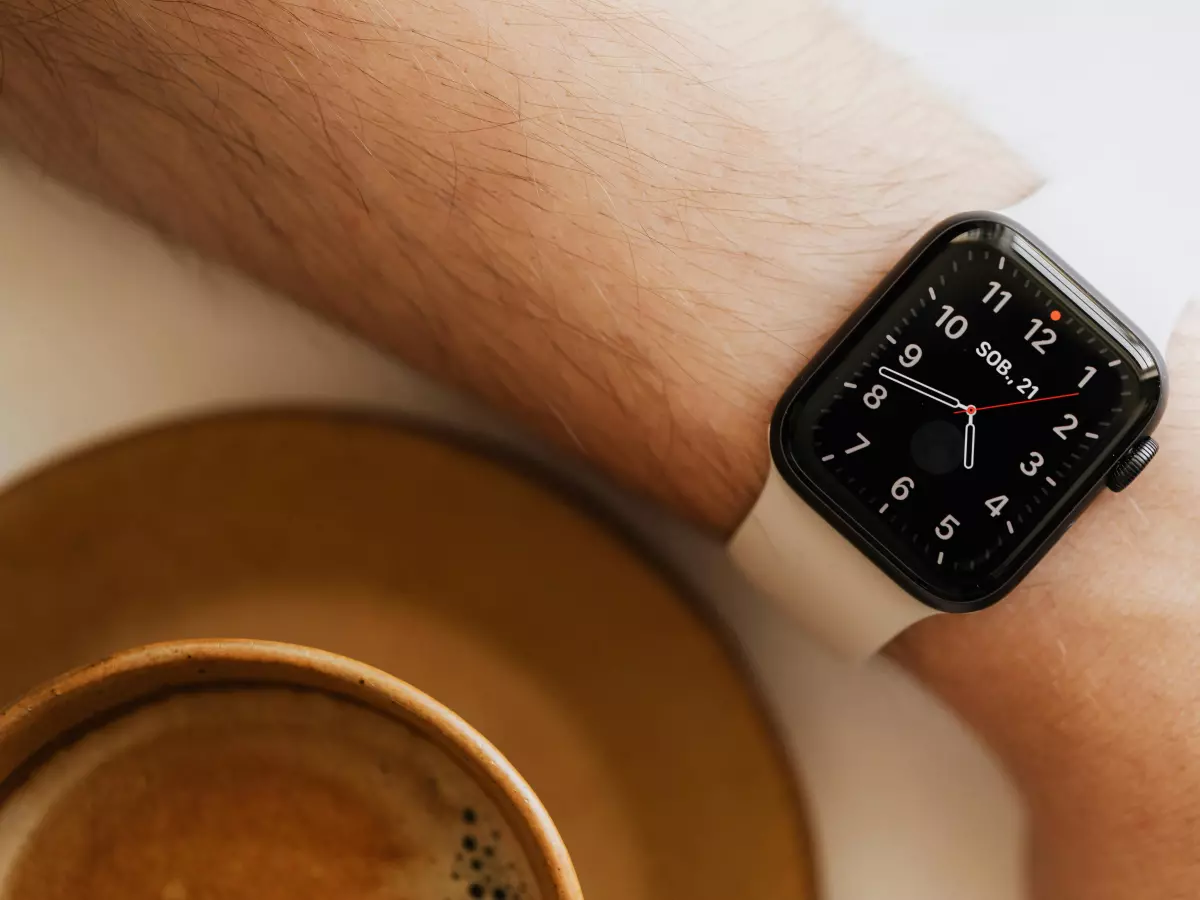 A close-up of a man's wrist wearing a modern smartwatch, showcasing the black watch face and its digital display. The watch is worn on a white band, and the background is a blurry image of a coffee cup.