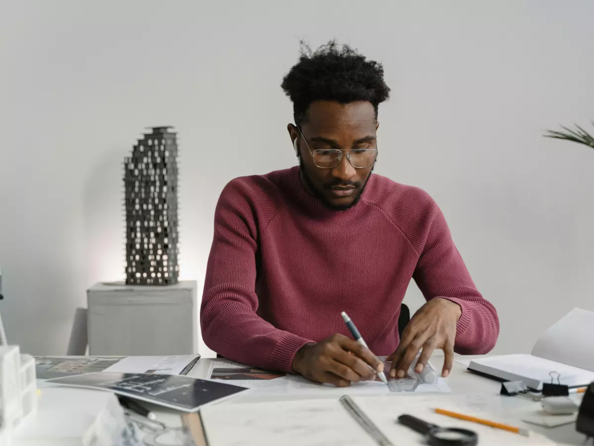 A person is sitting at a desk designing a PC. They're drawing diagrams and working with tools, with a laptop and plant in the background.