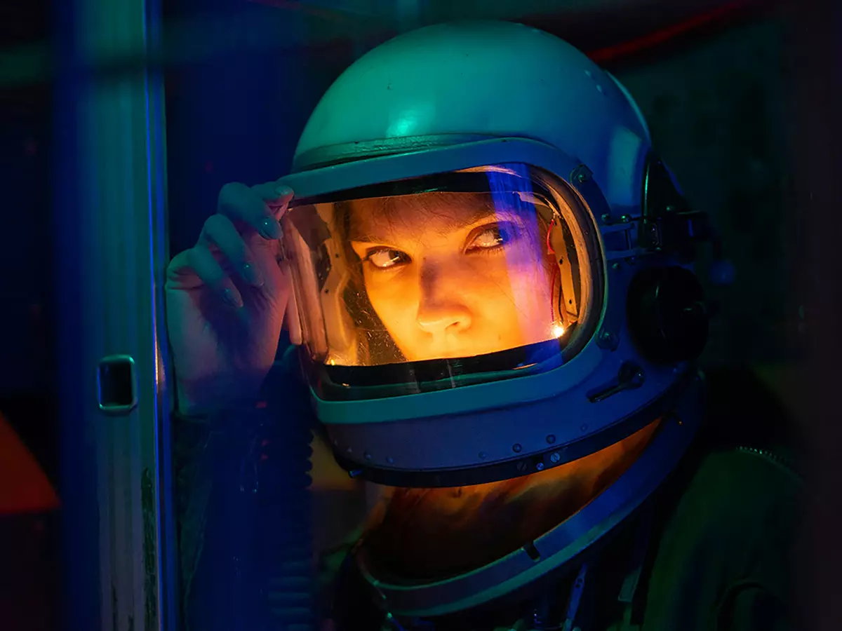 Close-up of a person wearing a futuristic astronaut helmet, illuminated with vibrant teal and orange light.