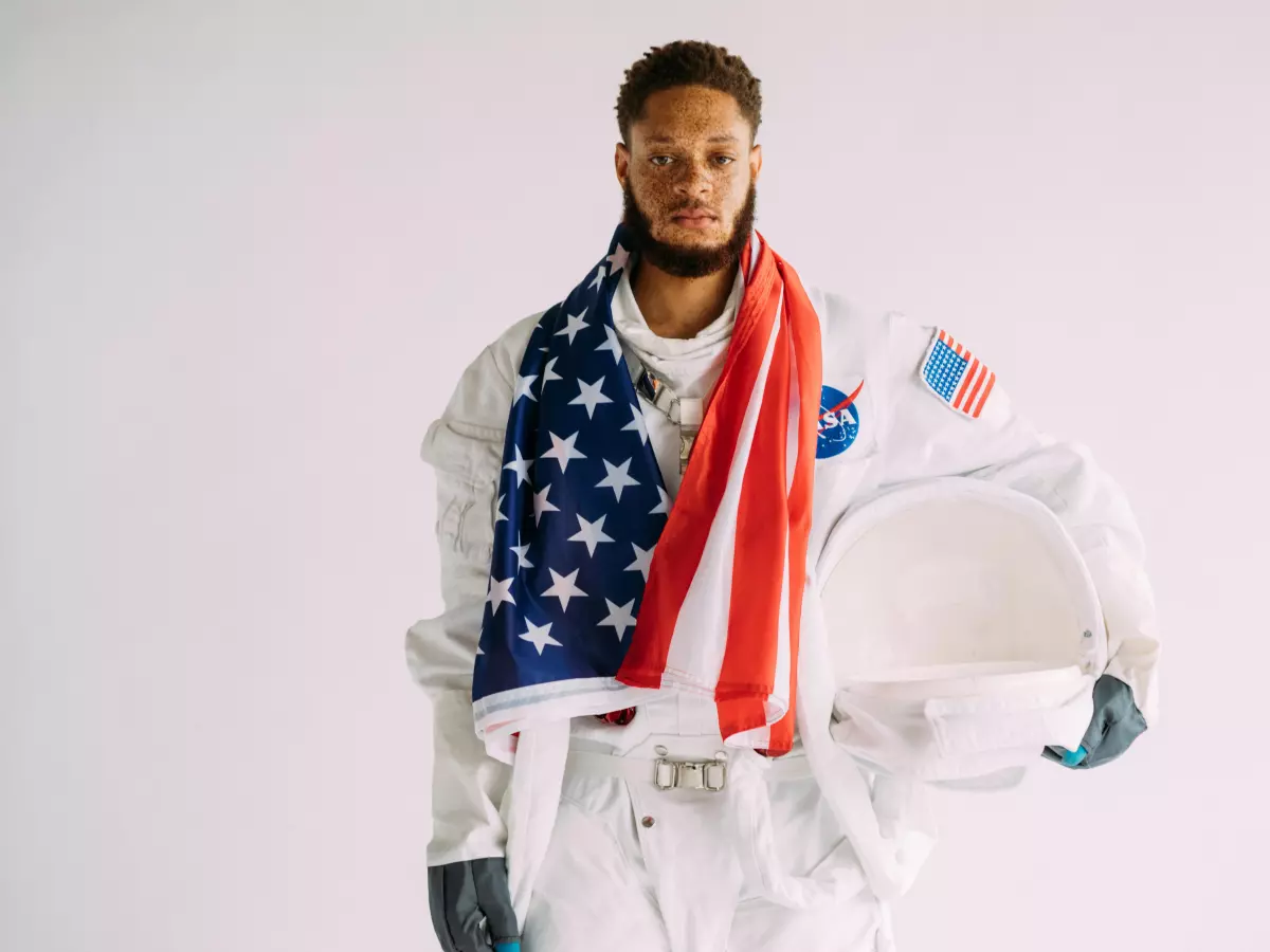 An astronaut wearing a white spacesuit and holding a US flag stands with a serious expression.