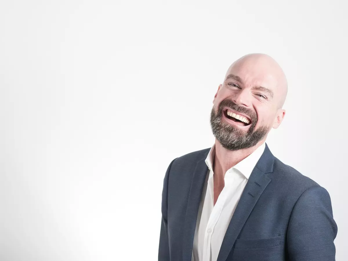 A bald man with a beard, wearing a suit, laughs brightly at the camera. He is in a studio setting with a white backdrop.