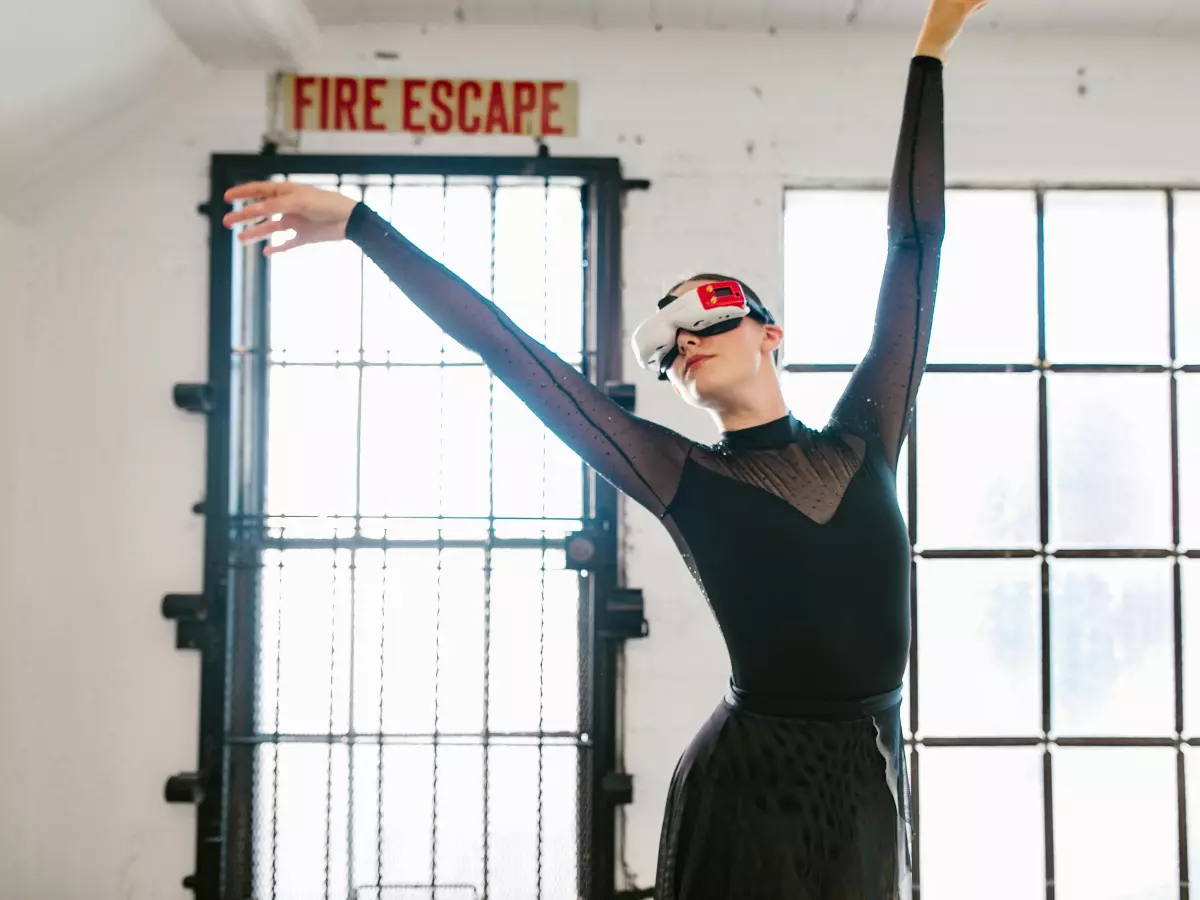 A woman wearing a VR headset is standing in a white room with windows, she has her arms raised. 