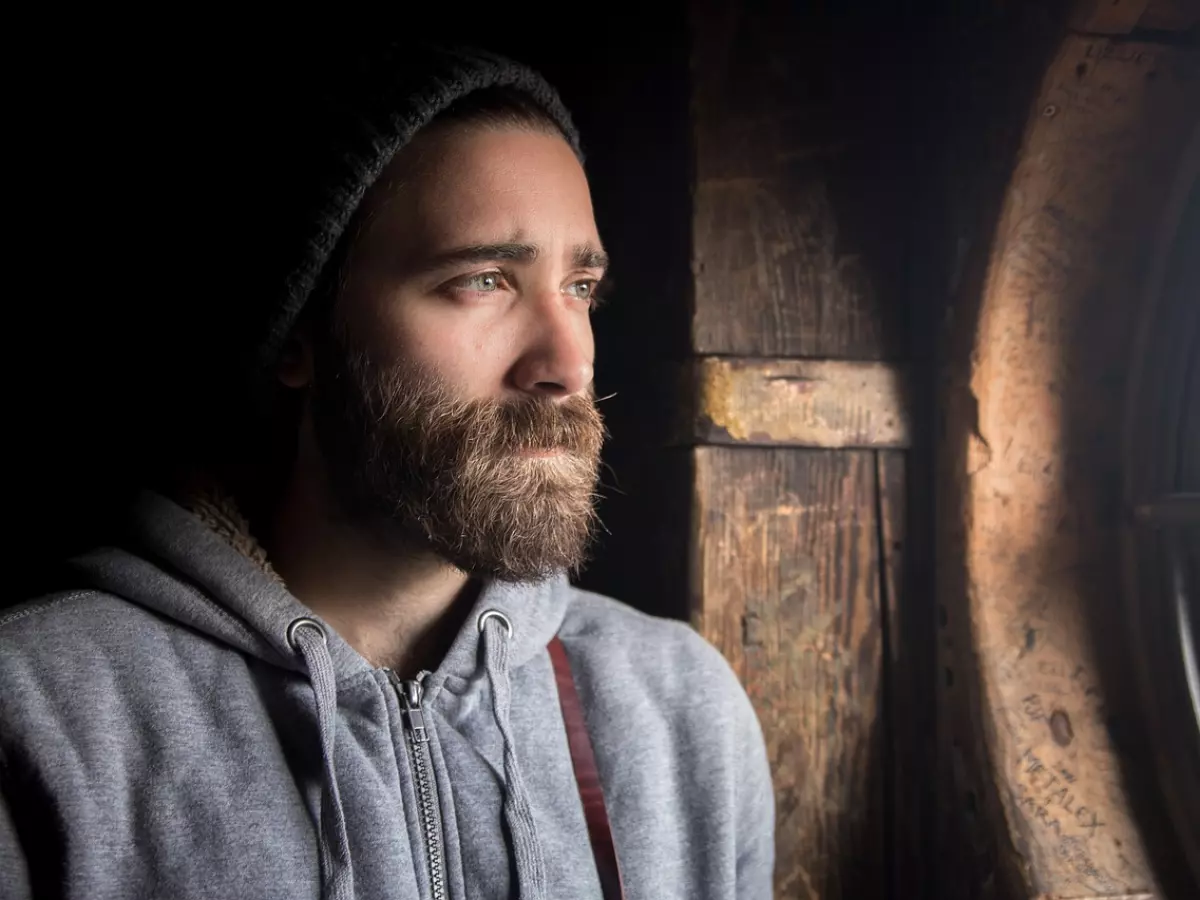 A man with a beard, looking out of a window.