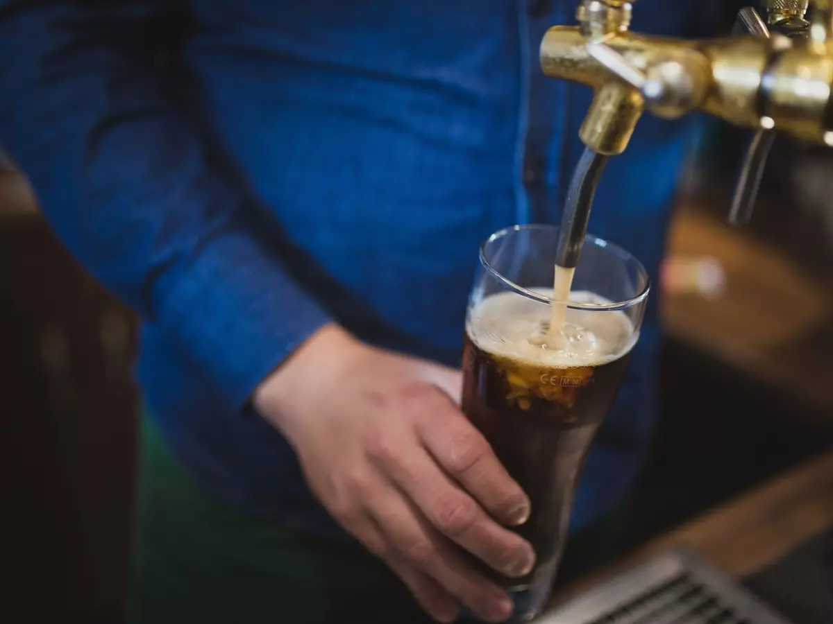 A person's hand pouring a beer from a tap into a glass. The person is wearing a blue shirt and the background is blurry.