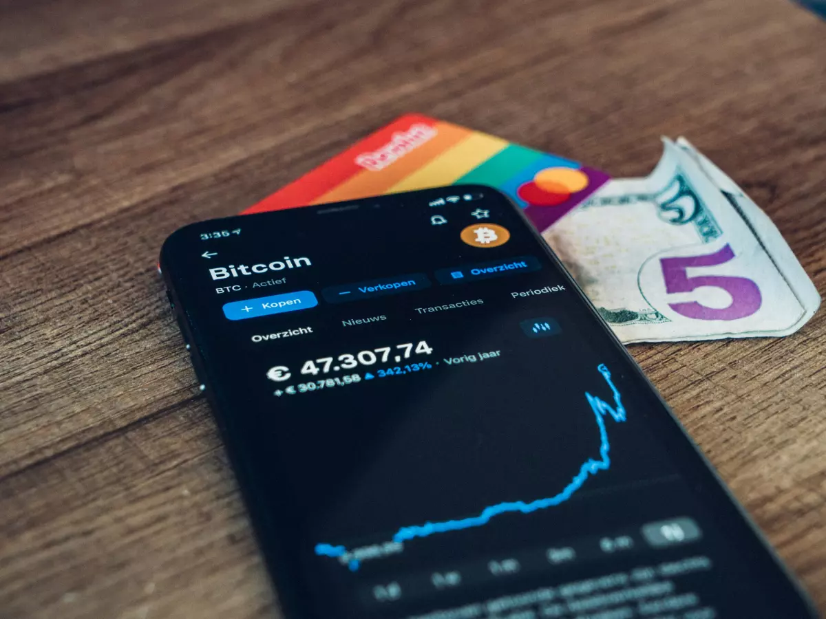 A smartphone displaying a Bitcoin chart, a credit card and a US dollar bill on a wooden surface.