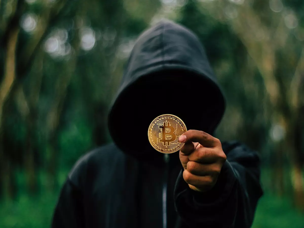 A hooded figure holding a Bitcoin coin, with a blurred background of forest.