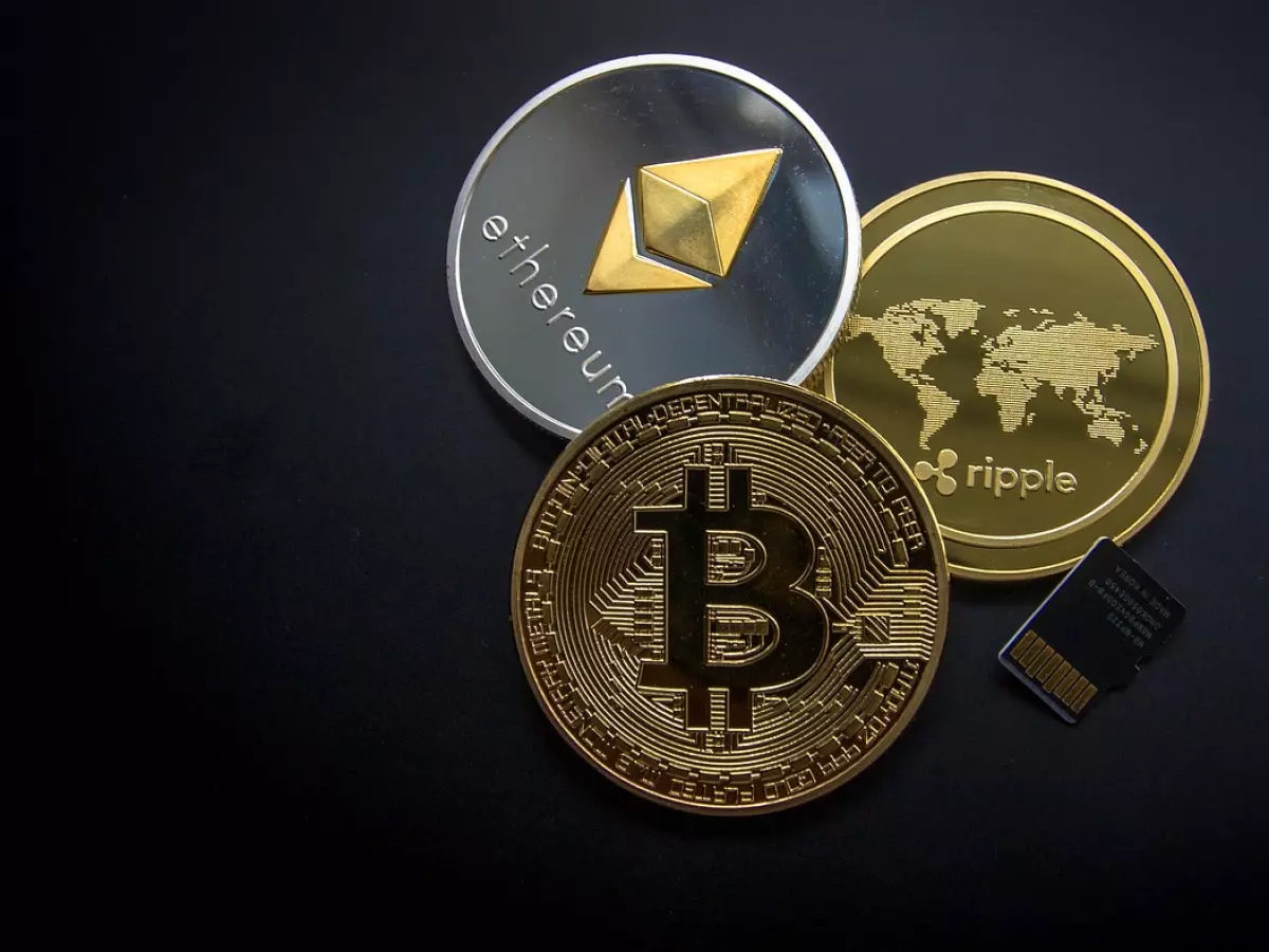 A photo of three cryptocurrency coins, Bitcoin, Ethereum, and Ripple, arranged on a dark background. A memory card is visible to the bottom right of the frame.