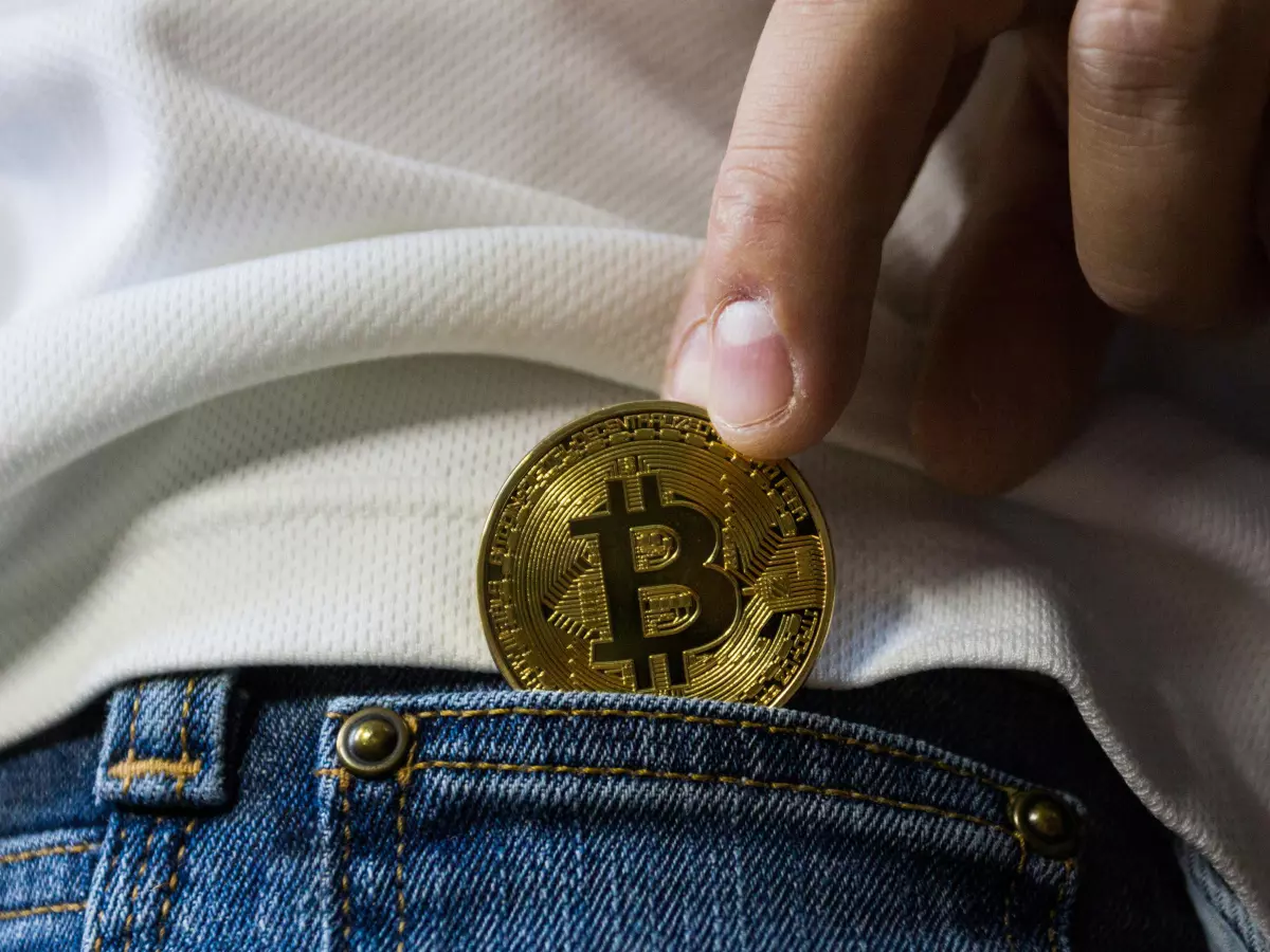 A close-up of a person's hand holding a Bitcoin coin, which is being placed in a pocket. The image is taken from a low angle, making the person's hand seem larger and more powerful.