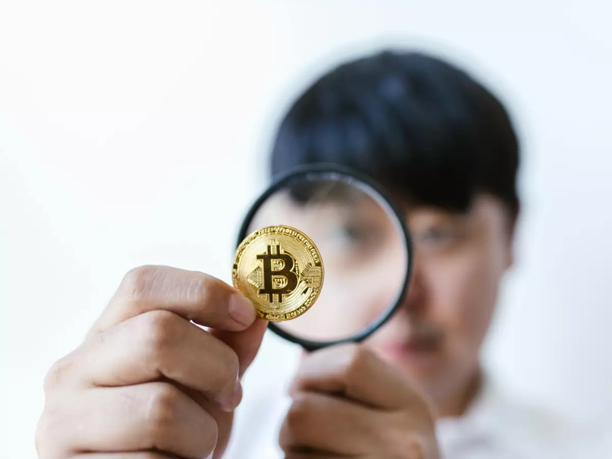 A close-up shot of a hand holding a Bitcoin coin with a magnifying glass in front of it.