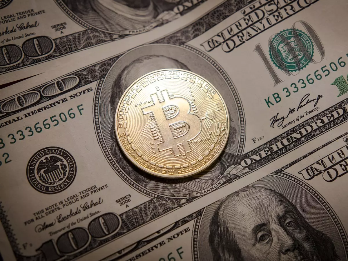 A gold-colored Bitcoin coin rests on top of two $100 US banknotes, capturing the visual representation of digital currency versus fiat currency.