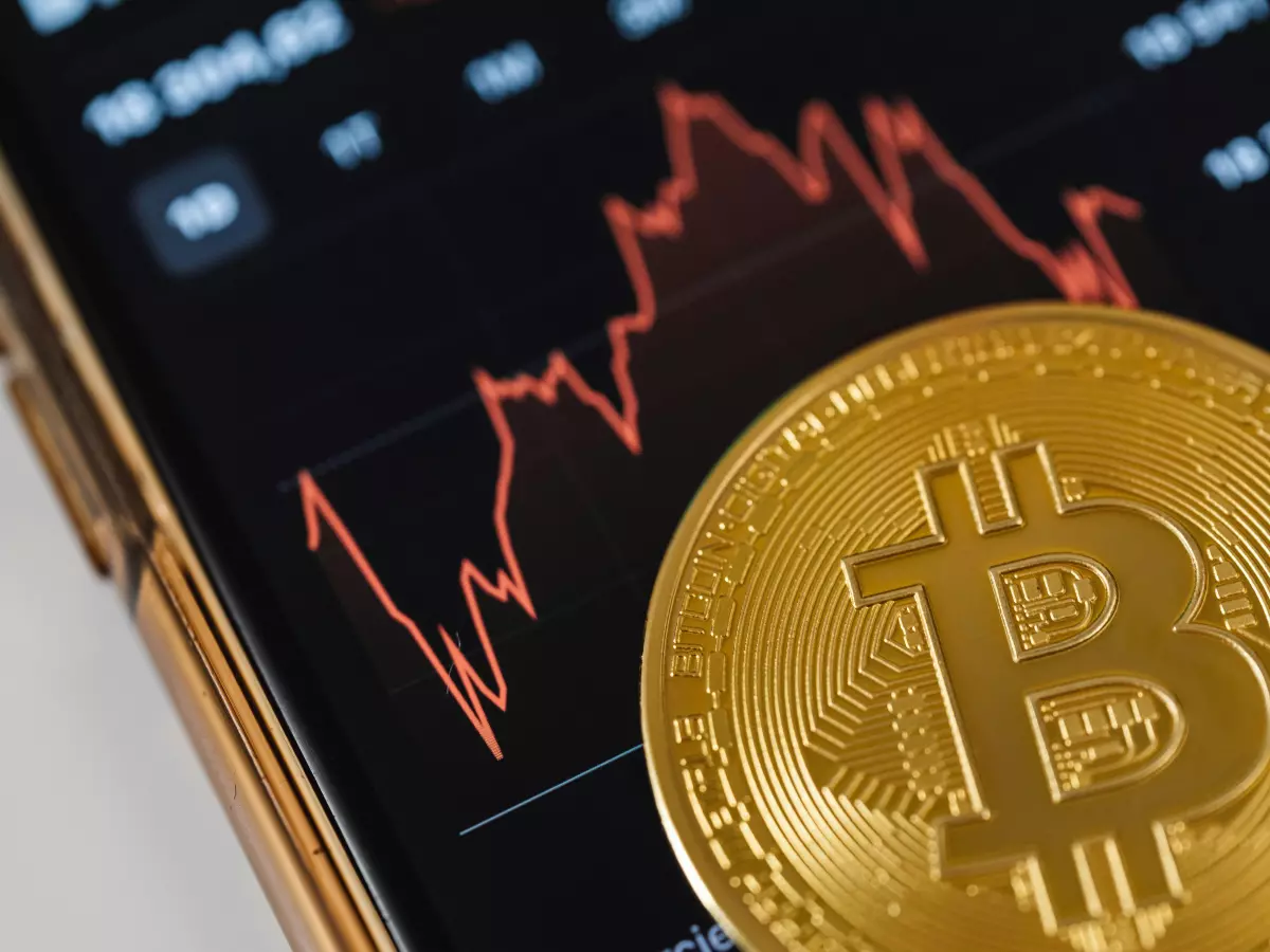 A gold Bitcoin coin rests on a smartphone screen displaying a red, dropping line graph. 