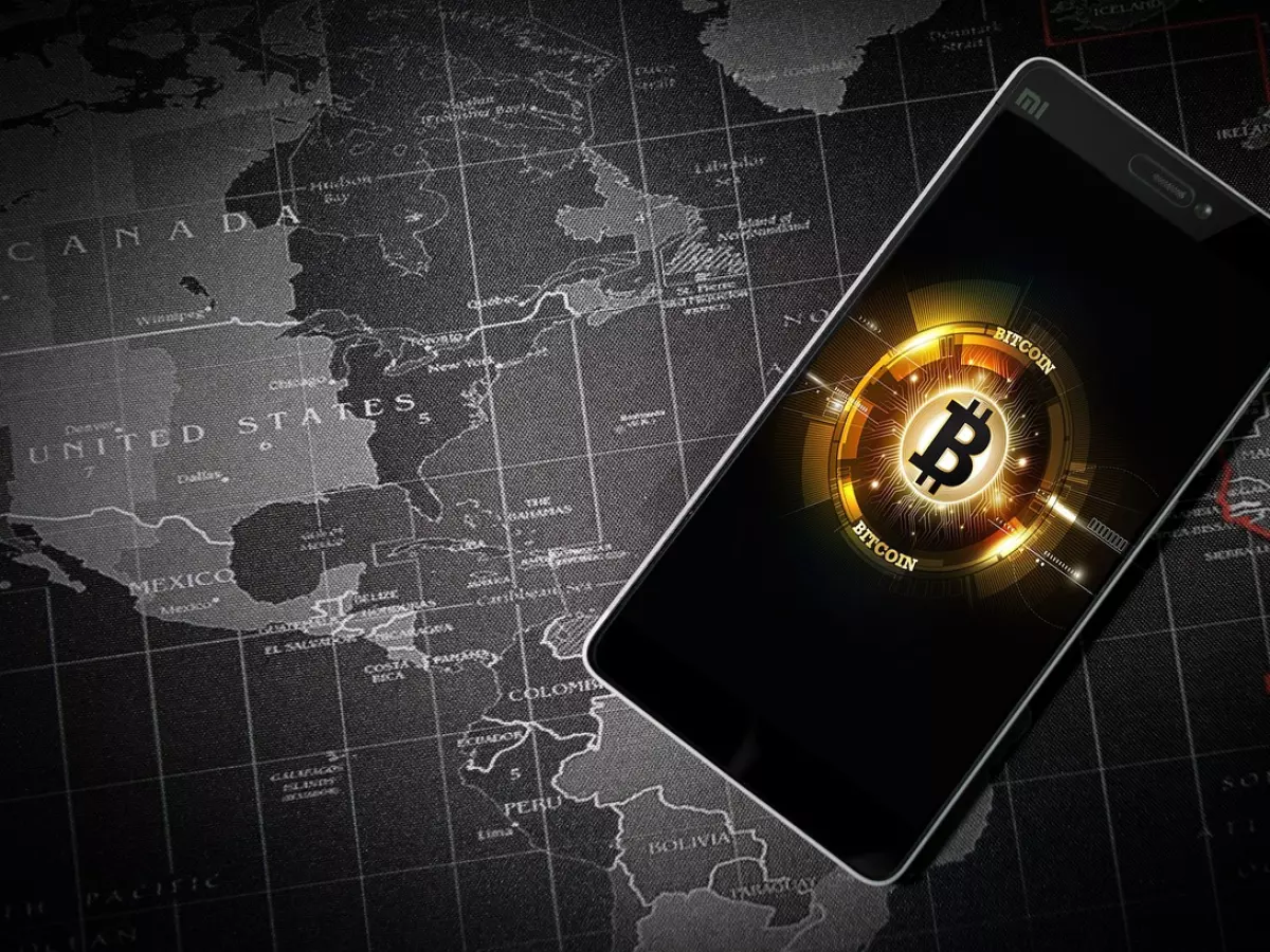 A smartphone with a Bitcoin logo on the screen is placed on top of a world map. 