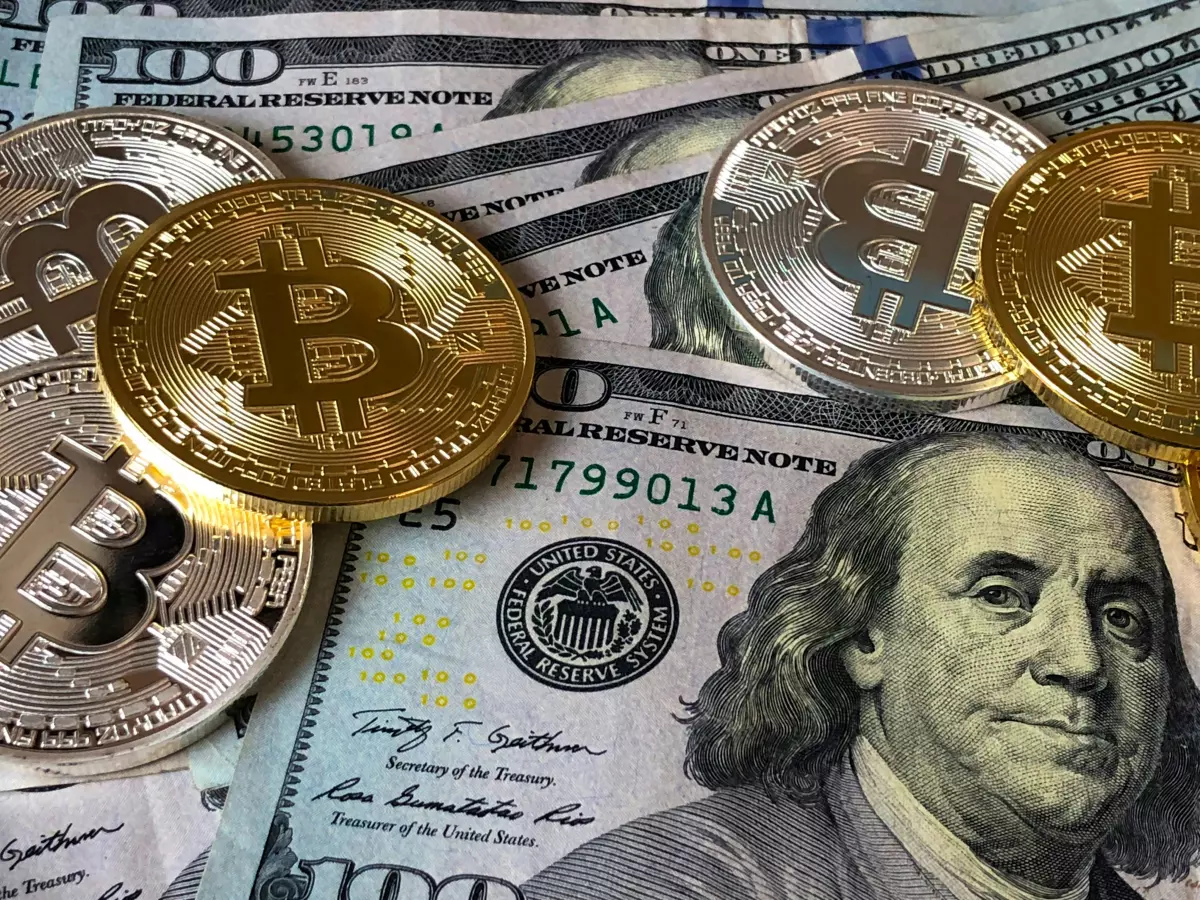 A close-up shot of Bitcoin coins on a bed of US dollar bills, with the face of President George Washington visible in the background.