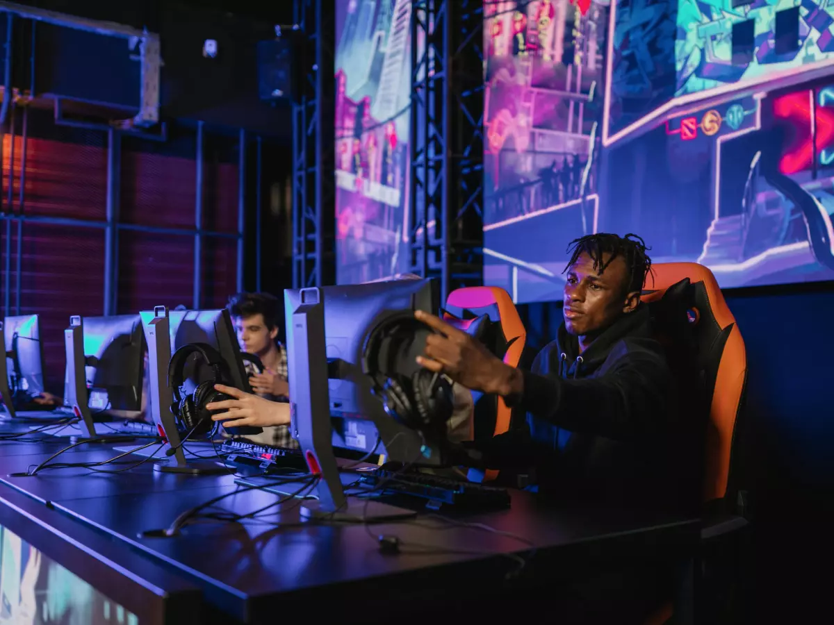 A young man in a gaming chair sits in front of a computer playing a game. The room is lit with neon lights and has a gaming vibe.